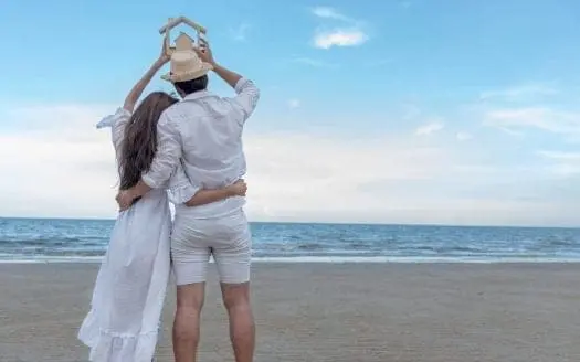 Un couple uni tenant une maquette de maison en bois au-dessus de leurs têtes sur une plage tranquille, symbolisant la construction d'un avenir commun et l'investissement immobilier après la signature d'un PACS