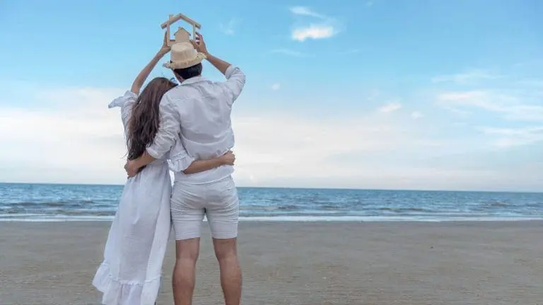 Un couple uni tenant une maquette de maison en bois au-dessus de leurs têtes sur une plage tranquille, symbolisant la construction d'un avenir commun et l'investissement immobilier après la signature d'un PACS